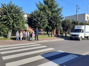 policjant z uczniami stoi przed przejściem dla pieszych. W tle budynki i samochód.