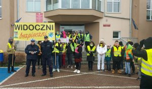 Policjanci, uczestnicy wydarzenia ubrani w kamizelki odblaskowe z banerami dotyczącymi promocji bezpieczeństwa na drodze stoją przed budynkiem szkoły.