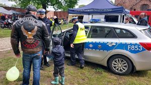 Stoisko promocyjne policji na pikniku motocyklowym. Policjant stoi obok radiowozu z uczestnikami wydarzenia.