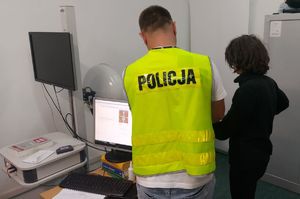 Policjant w kamizelce stoi tyłem z zatrzymana podczas daktyloskopii.