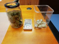 na stole stoi słoik w którym znajdują się zawiniątka z marihuaną obok leży waga elektroniczna oraz plastikowy pojemnik, w środku również zawiniątka z marihuaną