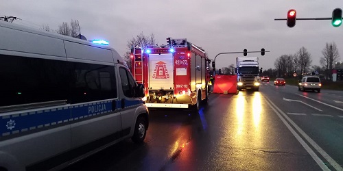 Droga , po prawej strony widać sygnalizację świetlną z zapalonym czerwonym i zielonym światłem, po lewej stronie stoi radiowóz oznakowany na sygnałach świetlnych, przed radiowozem stoi wóz strażacki, przed wozem strażackim parawan czerwony, z naprzeciwka stoi samochód ciężarowy.