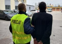 Dziedziniec Komendy Powiatowej Policji w Kutnie, w tle zaparkowane radiowozy, policjant w kamizelce odblaskowej z napisem Policja prowadzi zatrzymanego do radiowozu.