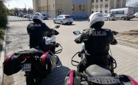 Policjanci siedzą na motocyklach, mają założone kaski. Na plecach widać napis Policja. Przed nimi widać budynek komendy i bramę wyjazdową