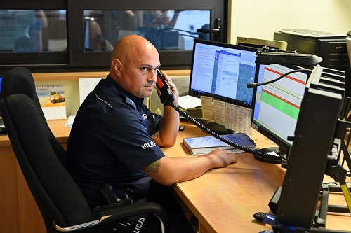 Policjant siedzi za biurkiem, trzyma w ręku słuchawkę od telefonu. Przed dyżurnym widać monitory ekranu