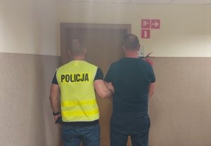 policjant w kamizelce odblaskowej z napisem Policja prowadzi korytarzem zatrzymanego
