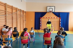 sala gimnastyczna dzieci siedzą w odstępach na krzesełkach policjantka prowadzi prelekcję