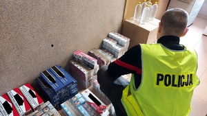 paczki papierosów poukładane na podłodze na korytarzu, obok pudełka kartonowe na nim stoją plastikowe butelki, przed nimi kuca policjant w kamizelce odblaskowej z napisem policja
