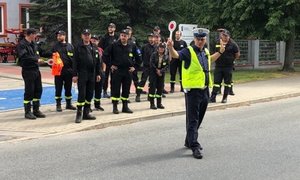 policjant stoi na drodze kieruje ruchem na chodniku stoją strażacy OSP