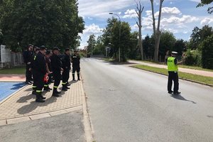 policjant stoi na drodze kieruje ruchem, strażacy OSP stoją na chodniku