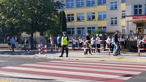 Przejście dla pieszych przy szkole podstawowej po drugiej stronie stoi policjant, przy szkolestoją dzieci
