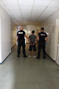 policjanci w mundurach z zatrzymanym stoją na korytarzu, mężczyzna ma z tyłu kajdanki.