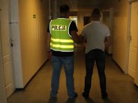 policjant Wydziału Kryminalnego w kamizelce z napisem Policja prowadzi zatrzymanego