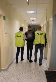 policjanci z Wydziału Kryminalnego w kamizelkach z napisem policja prowadzą korytarzem zatrzymanego, podejrzanego o uszkodzenie mienia