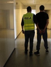 Policjant ubrany w kamizelkę odblaskową prowadzi korytarzem zatrzymanego mężczyznę.