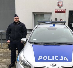 starszy aspirant Kotus stoi przy radiowozie, w tle Posterunek Policji w Nieborowie.