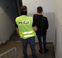 nieumundurowany policjant w kamizelce z napisem Policja prowadzi zatrzymanego po korytarzu w budynku komendy
