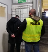 policjant  nieumundurowany w kamizelce z napisem policja prowadzi zatrzymanego po korytarzu budynku wieluńskiej komendy