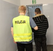 nieumundurowany policjant w kamizelce z napisem policja prowadzi zatrzymanego po korytarzu w komendzie policji