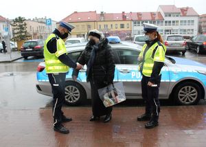 policjant i policjantka ruchu drogowego na tle radiowozu wręczają element odblaskowy pieszej