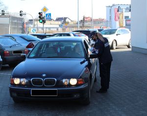 policjantka na parkingu wręcza kierowcy siedzącemu w aucie elementy odblaskowe i zawieszki zapachowe przechodniom
