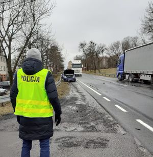 policjant w kamizelce z napisem Policja stoi przy drodze gdzie stoją samochód osobowy i ciężarowy które uczestniczyły w wypadku