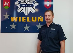Umundurowany policjant pozuje do zdjęcia na tle tablicy przedstawiającej gwiazdę policyjna z napisem Wieluń oraz herby gminy i powiatu wieluńskiego.