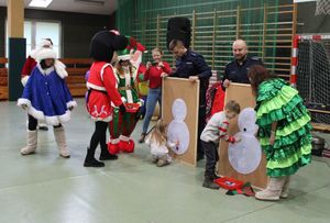 Elf, śnieżynka, Myszka Miki  stoją na sali gimnastycznej. Policjanci trzymają w rękach tablice, na których dziecko przypina kolejne elementy bałwana śnieżnego.