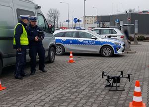 Dwaj policjanci stoją na parkingu,  jeden z nich obsługuje drona.