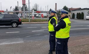 Dwie policjantki stoją przy drodze i nadzorują ruch , z boku częściowo widoczny radiowóz.