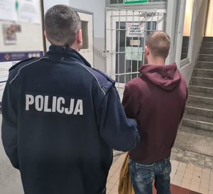 Policjant prowadzi zatrzymanego w kajdankach po korytarzu w budynku komendy.