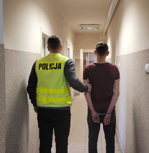 Policjant w kamizelce z napisem Policja prowadzi zatrzymanego po korytarzu.