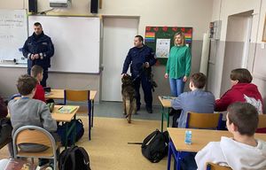 Policjant, wraz z przewodnikiem psa i jego podopiecznym stoją w sali lekcyjnej, w ławkach siedzą dzieci.