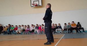 Policjant stoi w sali gimnastycznej na której na ławkach siedzą dzieci i prowadzi pogadankę.