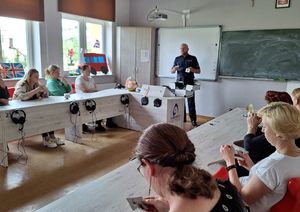 policjant w sali lekcyjnej w szkole stoi i prezentuje atrapy narkotyków i przedmiotów służących do ich przechowywania z walizki edukacyjnej . Wokół sali w ławkach siedzą rodzice i oglądają prezentowane przedmioty.
