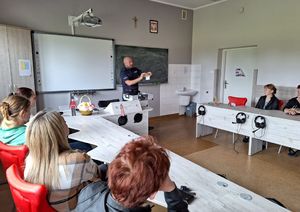 Policjant w sali lekcyjnej w szkole stoi i prezentuje atrapy narkotyków i przedmiotów służących do ich przechowywania z walizki edukacyjnej. W sali w ławkach siedzą rodzice.