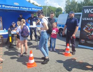 Stoisko profilaktyczne wystawione przez policjantów z wieluńskiej komendy na turnieju charytatywnym. Wokół uczestnicy imprezy. Jeden uczestnik pokonuje w narkogoglach tor przeszkód.