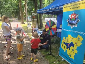 Stoisko profilaktyczne wystawione przez policjantów z wieluńskiej komendy na turnieju charytatywnym. Pod namiotem stoją policjanci oraz opiekunowie i dzieci biorące udział w konkursie wiedzy o bezpieczeństwie.