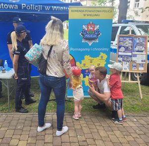 Stoisko profilaktyczne wystawione przez policjantów z wieluńskiej komendy na turnieju charytatywnym. Przy namiocie stoi policjantka oraz opiekunowie i dzieci biorące udział w konkursie wiedzy o bezpieczeństwie.