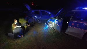 Rozbity samochód i radiowóz obok na trawie siedzi dwóch mężczyzn.