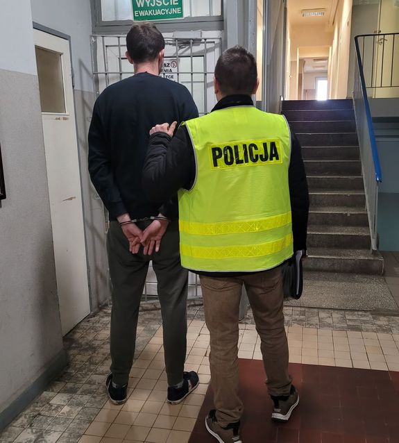 Policjant prowadzi zatrzymanego do drzwi wyjściowych w budynku komendy.