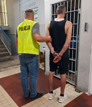 Policjant w kamizelce odblaskowej z napisem policja prowadzi zatrzymanego po korytarzu w budynku komendy.