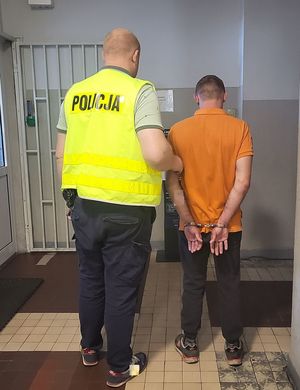 Policjant prowadzi zatrzymanego po korytarzu w budynku komendy.