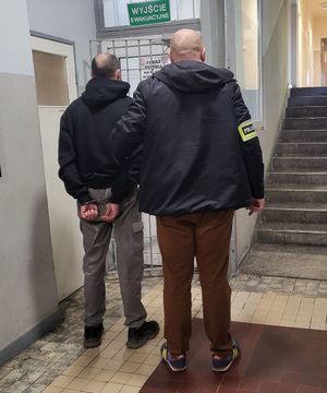 Policjant prowadzi zatrzymanego po korytarzu w budynku komendy.