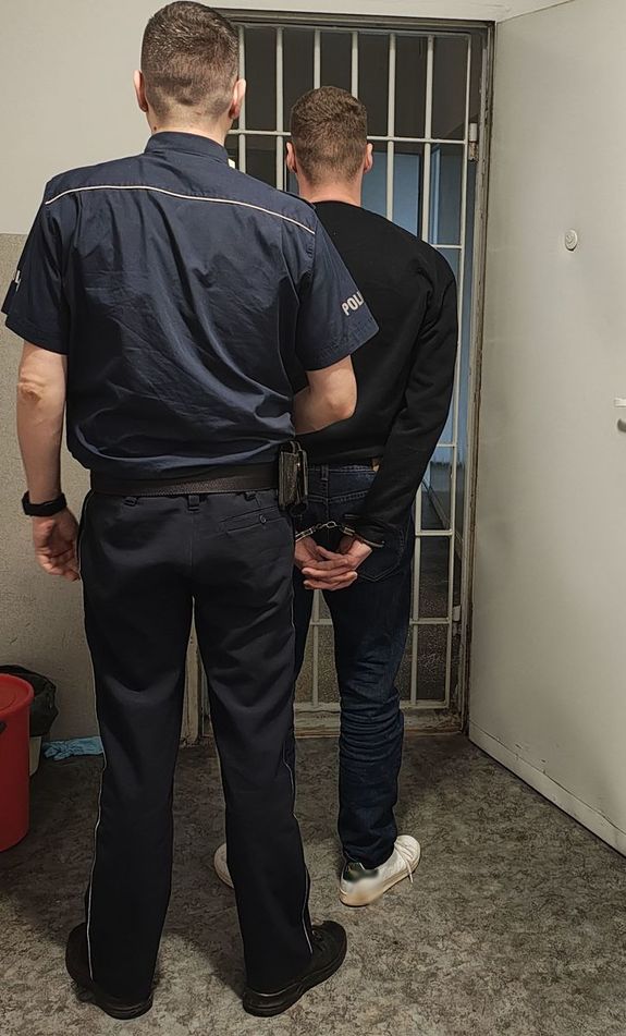 Policjant prowadzi zatrzymanego do pomieszczenia dla osób zatrzymanych.