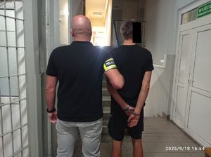 Policjant prowadzi zatrzymanego po korytarzu w budynku komendy.