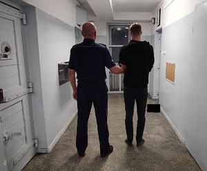 Policjant prowadzi zatrzymanego po korytarzu w pomieszczeniu dla osób zatrzymanych.