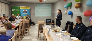 Policjantka w sali ośrodka kultury prowadzi prelekcję z seniorami, którzy siedzą przy stołach.