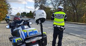 Policjant mierzy prędkość poruszających się pojazdów, obok stoi motocykl policyjny.