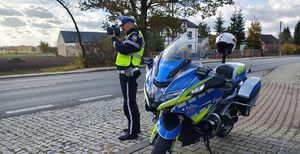 Policjant mierzy prędkość poruszających się pojazdów, obok stoi motocykl policyjny.
